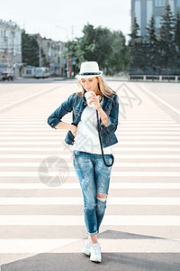 漂亮的轻女子戴着帽子牛仔裤夹克,带着次咖啡杯,站马路上走着斑马线,喝着咖啡,城市背景下跳舞图片