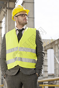 轻的男建筑师穿着保护工作服,双手站建筑工地的口袋里图片