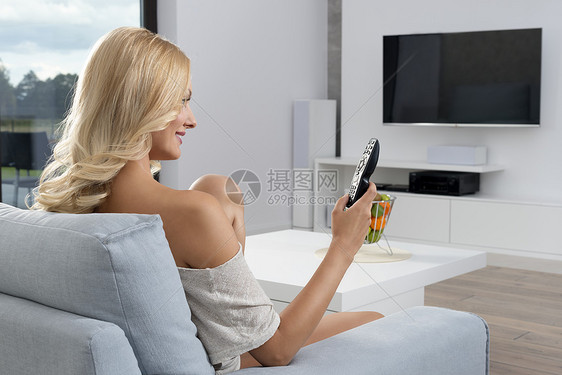 漂亮的轻女人坐电视前的蓝色沙发上她穿着舒适的毛衣,她头长长的金发,卷曲的头发图片