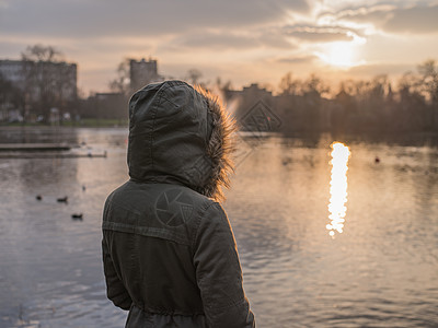 个穿着厚厚的保暖外套的人站公园的池塘边,正冬天看日落图片