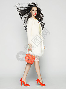 美丽的黑发模特穿着时尚服装工作室摆姿势穿着外套,手提包,红鞋图片