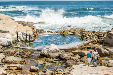 两个人站海角镇的岩石上,附近个人了潮汐池,看着海浪撞击岸边的岩石图片