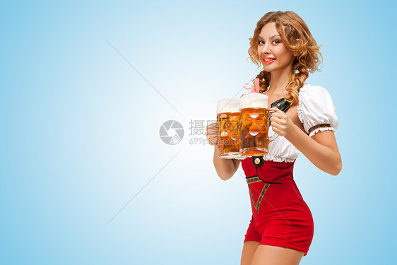 轻兴奋感的瑞士女人穿着红色跳线短裤吊带的传统Dirndl,蓝色背景上提供两个啤酒杯图片