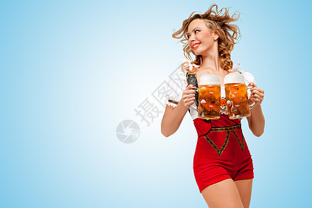 轻的微笑感的瑞士女人穿着红色毛衣短裤,吊带的形式传统的Dirndl,着两个啤酒杯,边看着蓝色的背景图片