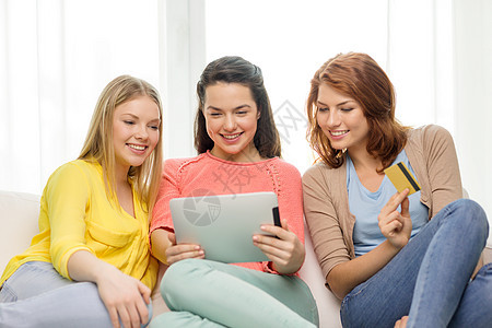 三个微笑的十几岁女孩与平板电脑信用卡在家里图片
