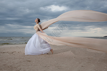 穿着米色长裙的女模特海滩上摆出动感十足的造型挥动布料飘动的长裙背景图片