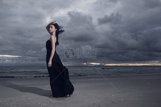 美丽的黑发模特穿着黑色长裙站海滩上多云天气日落图片