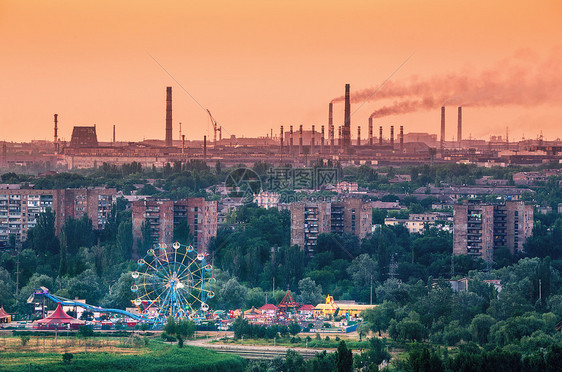 带摩天轮的游乐园日落时烟雾弥漫的冶金工厂背景上的建筑物生态问题,大气污染物烟的管子重工业工厂钢铁厂图片