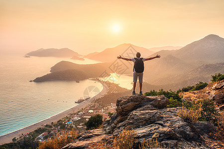 快乐的运动男人,带着背包,举手臂,站岩石上,夏天的日落时看着海岸山脉人,海,山脊橙色的天空奥卢德尼斯,火鸡背景图片