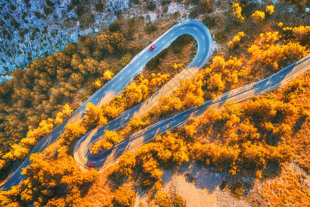 空中观看山曲线道路与汽车,橙色森林日落欧洲秋季景观与沥青道路,树木岩石上穿过公园的高速公路飞行无人机的顶部视图图片
