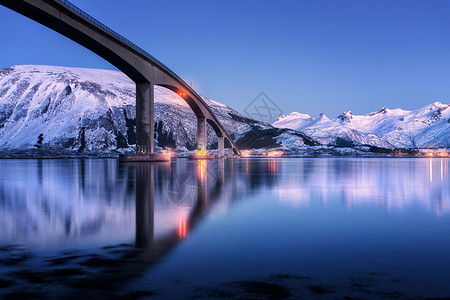 桥照明,雪覆盖着山,村庄蓝天,水丽的倒影夜间景观与桥梁,雪岩反射海上挪威洛福滕岛的冬天图片