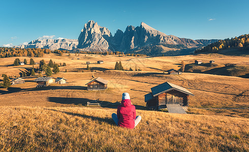 坐山上的女人秋天的日落时看着草地山脉阿尔佩迪西斯,白云石,意大利风景与女孩穿着红色夹克,田野与黄草,木屋岩石图片