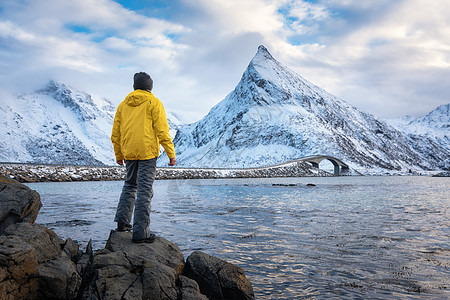 穿着黄色夹克的运动男子站海边的石头上,冬天的日落时,雪山多云的天空风景与旅行者,水,弗雷德旺桥洛弗顿群岛,挪威图片