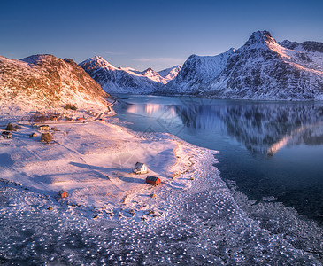 鸟瞰积雪覆盖的山脉,房屋,霜冻的海岸蓝天反射水日落冬季景观与海洋,雪岩,罗布,道路挪威洛福滕岛的个小村庄图片