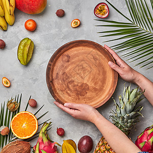 成熟的热带水果,椰子,芒果,杨桃,荔枝棕榈叶灰色的混凝土背景女人的手着个空的棕色圆盘子,个的副本平躺图片