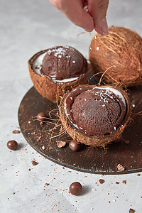 两个椰子半与冷冻新鲜棕色甜点整个椰子与女人的手个圆形木板上的灰色石桌与夏季饮食女手椰子洒巧克力冰淇图片
