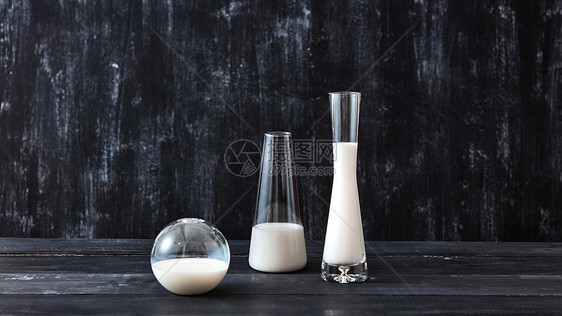 璃锥球形的容器,黑色背景上的深色木桌上装满白色液体,并放置文字犹太节日的三杯乳制品牛奶酸奶站黑色的木桌上图片