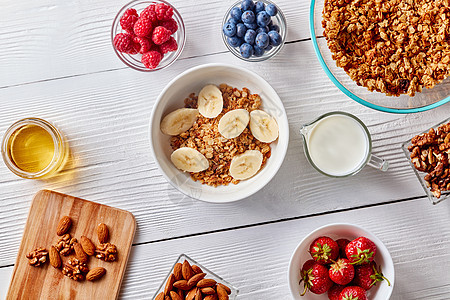 自制燕麦麦片,香蕉,坚果,浆果蜂蜜放璃碗里,牛奶放白色的木制背景上的风景健康的早餐成分与自制的麦图片