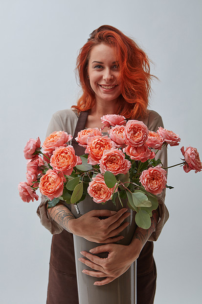 个微笑的红发女人穿着棕色围裙,着个花瓶,2019的玫瑰颜色,生活灰色背景上的珊瑚潘通,文字花店的图片
