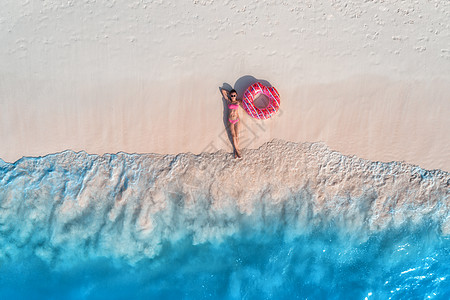粉红色沙滩鸟瞰美丽的轻躺着的女人,粉红色的甜甜圈游泳环白色的沙滩上,靠近大海,日落时波浪暑假苗条女孩的最高视野,清澈的蔚蓝的背景