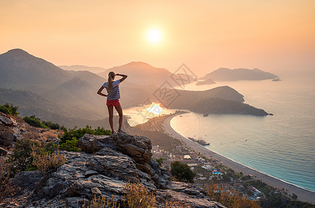 轻的女人站岩石的顶端,看着海滩山脉,夏天五颜六色的日落风景与女孩,海洋,山脊橙色天空与太阳奥卢德尼斯,火鸡图片