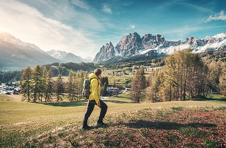 轻人穿着黄色夹克,带着背包,春天的日落时分,绿色的草地上靠山行走风景与运动的家伙,雪岩,树木,建筑物,蓝天意大利图片