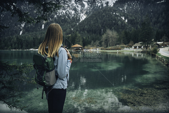 轻的女人带着背包站山湖的海岸上,春天的阴天意大利旅行风景与苗条的女孩,倒影水,雪岩,绿树复古色调带着背图片