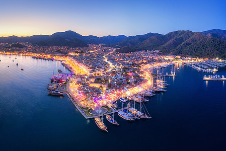 鸟瞰船只美丽的城市夜间马尔马里斯,土耳其全景景观与船码头湾,海洋,城市灯,山,蓝天黄昏无人机的顶部视图日落时图片