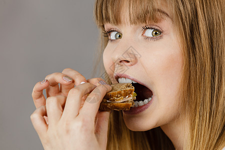 轻的女人吃三明治,用张开的嘴咬人食物,卡路里,节食的工作室拍摄灰色背景,侧写视图女人吃三明治,咬人图片
