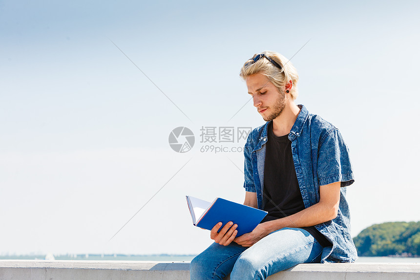 度假,户外休闲时间,内向放松的坐着的时髦男人穿着牛仔裤,穿着牛仔裤,阳光明媚的日子里外看书,背景大海阳光明图片
