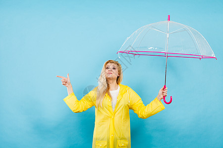 金发女人穿着黄色雨衣,着透明的雨伞,检查天气否下雨,用手指指着电脑图片