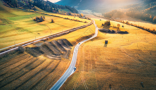 秋天日落时山间山谷道路的鸟瞰图沥青道路的顶部景观,铁路,带橙色草地的山丘,阳光,黄树,建筑物秋天的公路田野风景图片