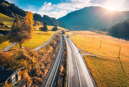 秋天日落时山间山谷道路的鸟瞰图沥青道路的顶部景观,铁路,带橙色草地的山丘,阳光,黄树,建筑物秋天的公路田野风景图片