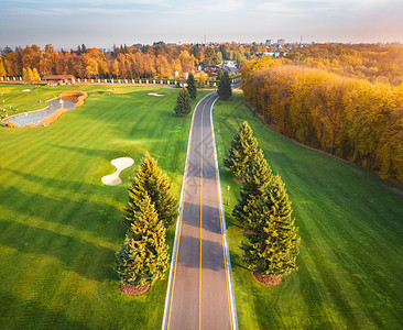 秋天日落时穿过高尔夫球场秋季巷道绿色高尔夫球场的鸟瞰图景观与路线,草地,绿色松树,橙色森林,美丽的天空看图片