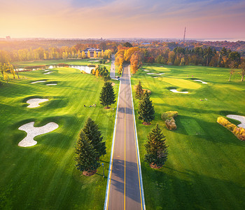 秋天日落时穿过高尔夫球场秋季巷道绿色高尔夫球场的鸟瞰图景观与路线,草地,绿色松树,橙色森林,美丽的天空看背景图片