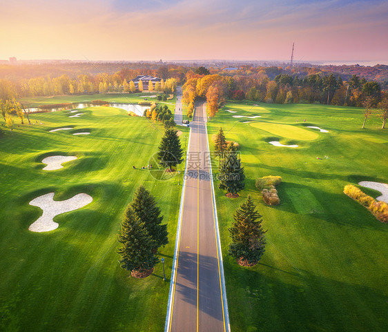 秋天日落时穿过高尔夫球场秋季巷道绿色高尔夫球场的鸟瞰图景观与路线,草地,绿色松树,橙色森林,美丽的天空看图片
