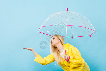 金发女人戴着黄色雨衣,着透明的雨伞检查天气,如果下雨的话图片