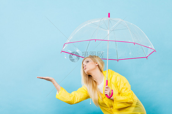 金发女人戴着黄色雨衣,着透明的雨伞检查天气,如果下雨的话图片