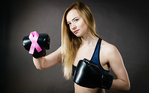 轻的运动女子戴拳击手套,带粉红色丝带,乳腺癌的象征与疾病搏斗,女的动机工作室拍摄黑暗的背景轻的女人戴着粉红色丝图片