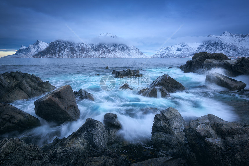 海滩上的石头模糊的波浪中,蓝天与低云雪山黄昏的冬天挪威洛福顿群岛的海岸戏剧的景观与海洋,岩石雾中喜怒无常的图片