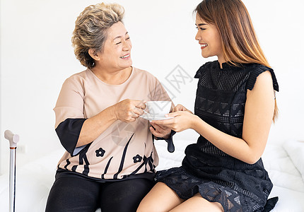 照顾者妇女给床上的老妇女喝咖啡,并家里检查她图片