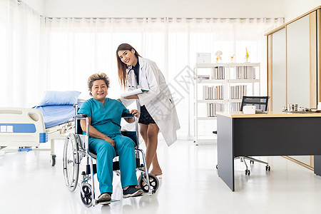 亚洲自信医生照顾者谈论健康问题与老妇女病人坐轮椅上的医院病房,而每天检查图片