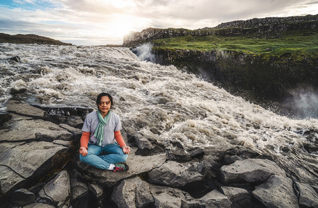 年轻女子坐在河边和大瀑布旁做冥想瑜伽图片