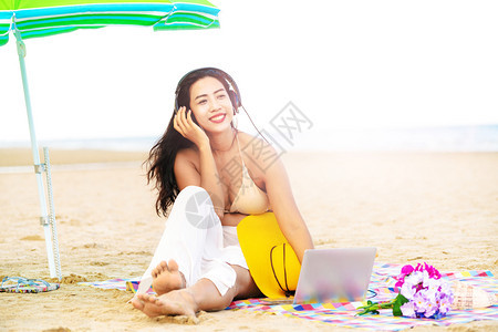 夏天坐在热带沙滩上戴着耳机听音乐的年轻快乐女子图片