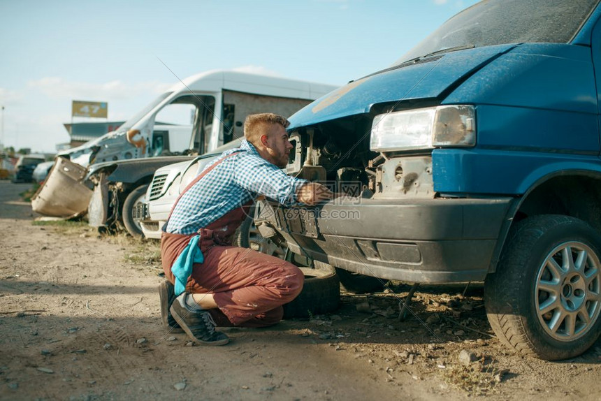 废弃汽车垃圾场里的一名男修车工将头卡在引擎盖下修理图片