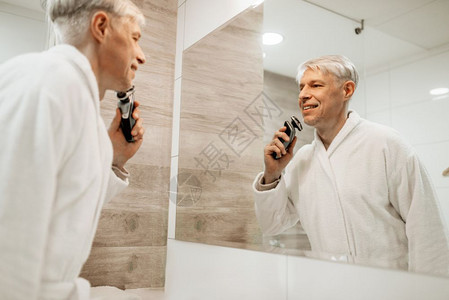穿着浴袍的快乐成年男子在浴室的镜上刮胡灰发成年男子带着电剃须刀快乐成年男子在浴室的镜上刮胡图片