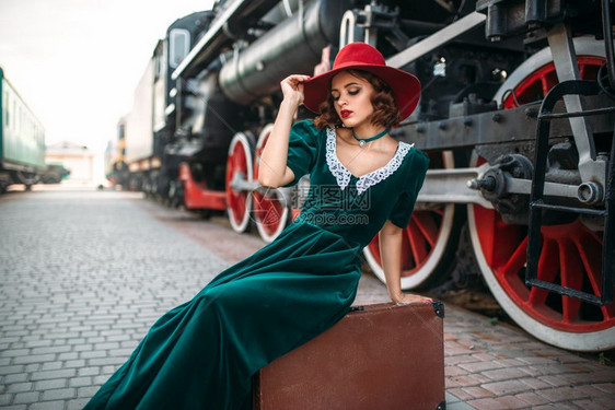红帽子的女士坐在手提箱上和旧式蒸汽列车对着旧式火头老铁路引擎古老的铁路车辆红帽子的女士跟旧式蒸汽列车对着图片