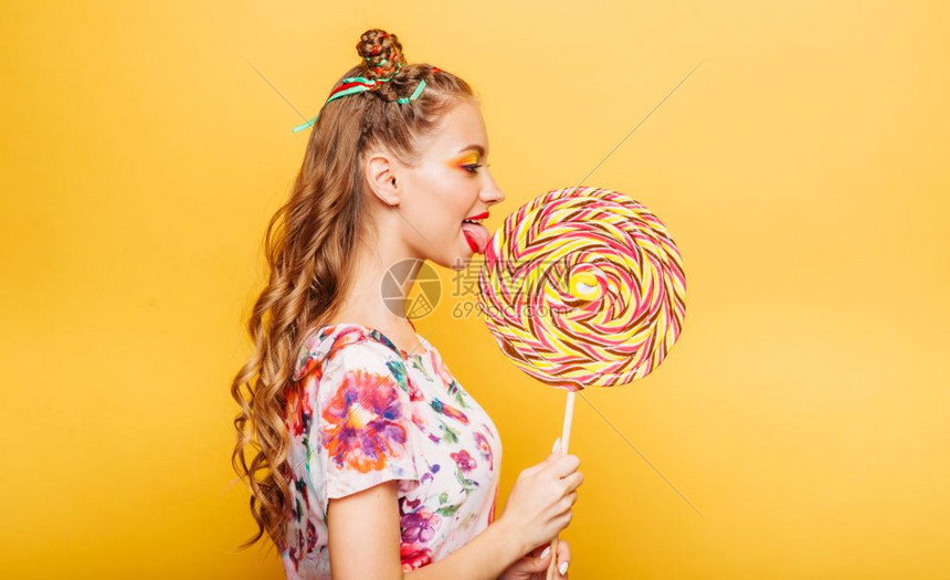 在黄色背景中美丽的年轻女孩笑脸吃着巨大的糖果图片