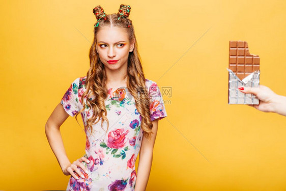美丽的年轻女士拒绝吃巧克力吧穿着金色卷发的明亮女孩穿着夏季彩色服装的时髦女孩背景上是黄色的墙女士拒绝吃巧克力吧图片