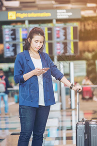 在国际机场使用智能电话等待报到时间的飞行信息委员会附近携带行李的年轻旅游女客图片
